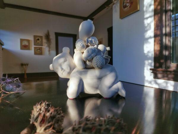 A photograph of a white ceramic sculpture of a small kneeling figure balancing fruits on its back.