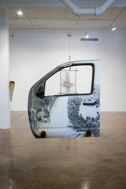 An installation photograph of a work by Andrei Renteria. A white truck door that has a graphite drawing on it hangs from a wire.