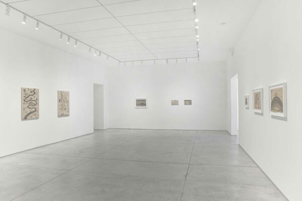 A photograph showing the interior of a white-walled art gallery. Drawings on canvas are hung on the walls of the gallery.