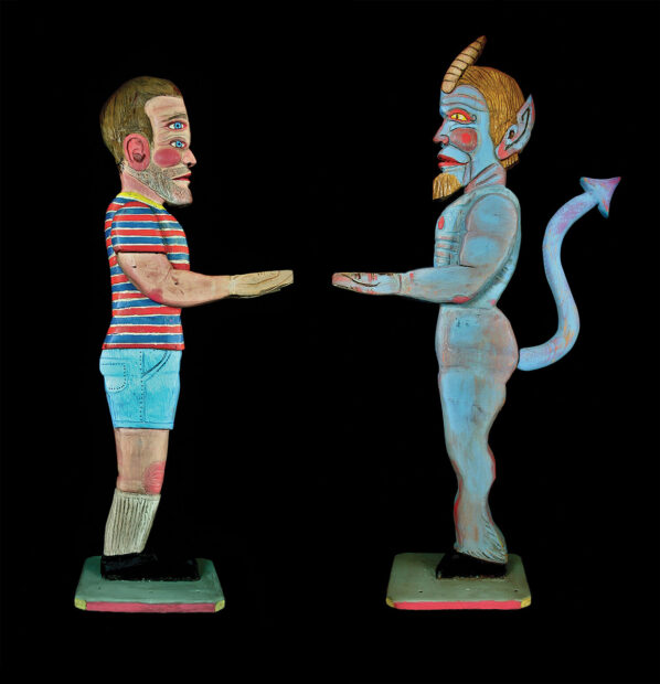 Two wooden sculptures of figures by Chuck & George. The figures are facing each other and one has been painted as a man with two sets of eyes who is wearing a t-shirt, shorts, knee-high socks and black shoes. The other figure is painted like a blue devil with hoofed feet and a pointed tail.