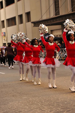 A photograph of a small group of dancers dressed in Mrs. Clause outfits as they march in the Dallas Holiday Parade.