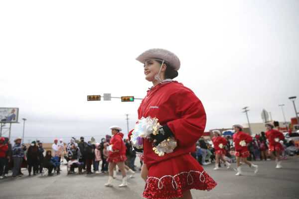 A photograph from the 2021 El Paso Sun Bowl Parade featuring a group of young dancers wearing red skirts, red jackets, white cowboy hats, and holding white pom poms.