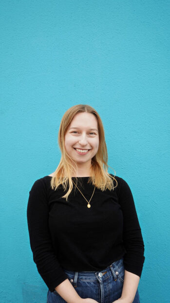 A photograph of Emily Fens set against a bright blue backdrop. She looks straight into the camera with a wide smile.