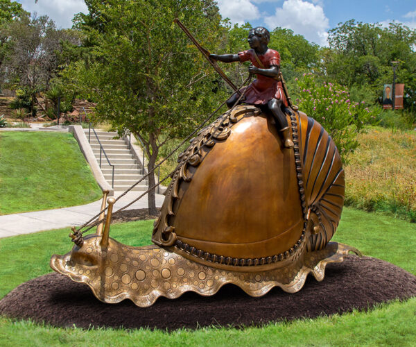 A photograph of a large-scale bronze sculpture of a young child riding atop a giant snail. Artwork by Hank Willis Thomas.