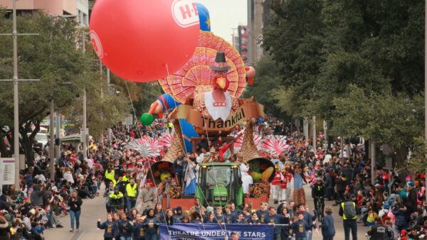 A photograph of a large parade float featuring a Turkey. The float is surrounded by people lining either sides of the street.