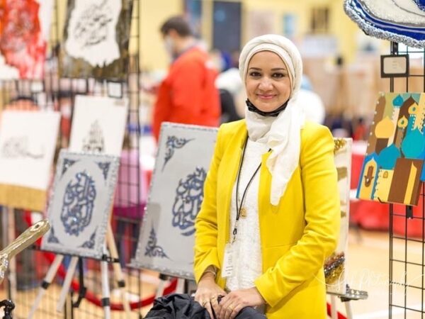 A photograph of an artist standing in front of her booth displaying calligraphy-based artwork. 