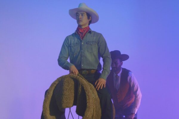 A still image from a video by Kenneth Tam. The image shows a cowboy sitting on a covered barrel as if it is a horse. Behind him lurks another cowboy.