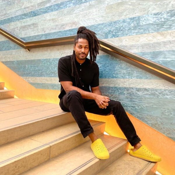 A photograph of Kenyon Adams sitting on a set of stairs at the Blanton Museum of Art. He looks straight into the camera with a relaxed expression and pose.