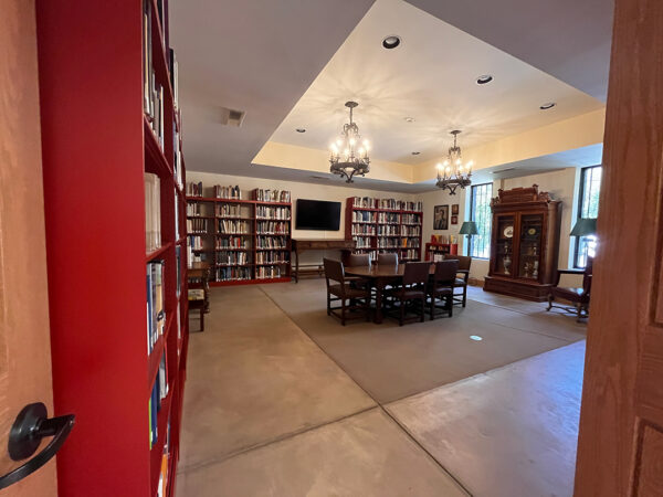 A photograph of the Old Jail Art Center library. Tall red bookshelves line the walls and a table with six chairs is in the center of the room. 
