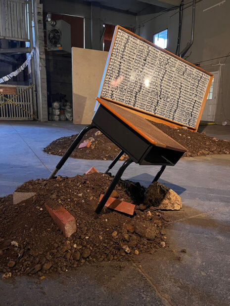 A photograph of an installation by Phallon Lauderback Wright which features a disheveled school desk and white board stuck in a pile of dirt and bricks. The white board and desk have repeated text written across their surfaces.