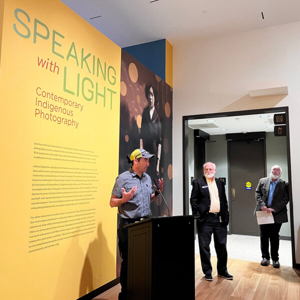 A photograph of Will Wilson standing at a podium and speaking about co-curating an exhibition with John Rohrbach, who stands in the background.