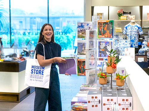 A photograph of a woman shopping at a museum store. She holds a tote bag that reads, "Museum Store Sunday. Be a patron."