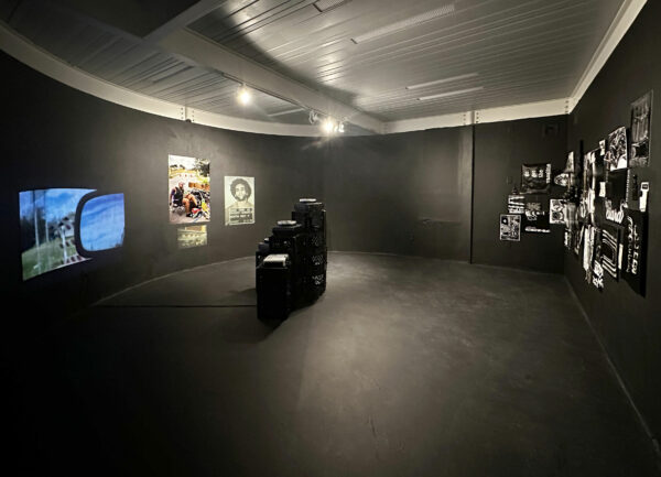 Installation view of speakers, video, and photos in a black room