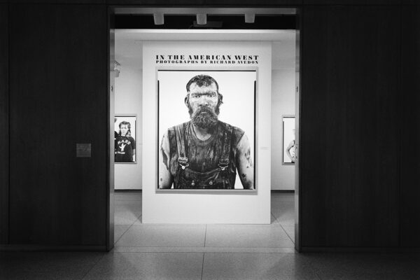 A black and white installation image from the 1985 exhibition "Richard Avedon: In the American West." The photo shows a large-scale black and white portrait framed by a darkened doorway.