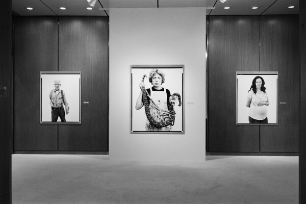 A black and white installation image from the 1985 exhibition "Richard Avedon: In the American West." The photo shows three black and white portraits hanging on a gallery wall.