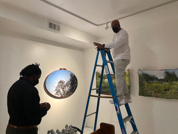 A photograph of Alfred Walker adjusting lighting in a gallery with an assistant helping sort track lights.