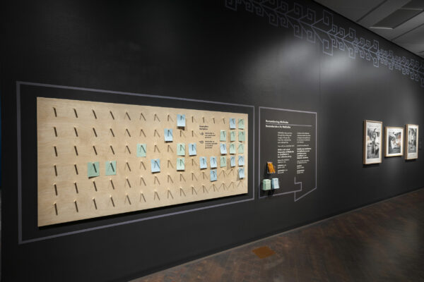 Sobre el muro negro de una galería se encuentra un tablero rectangular con clavijas. Debajo de algunas clavijas hay cuadrados de papel azul y verde.