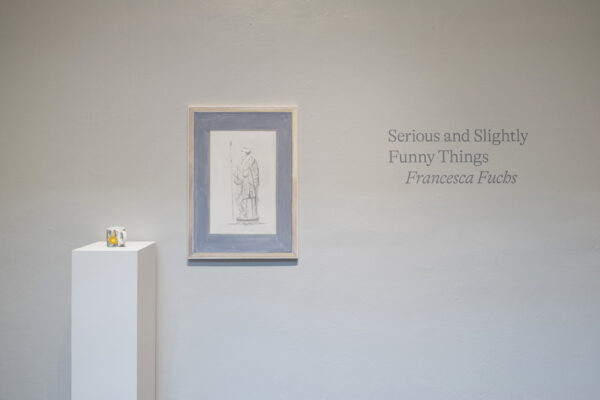 Title wall and installation view of a work on a wall and a cup on a pedestal