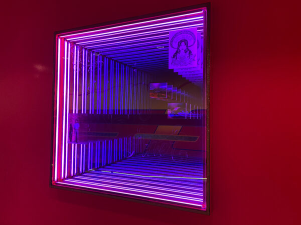 A photograph of a part of a larger installation by Guadalupe Rosales. The photograph shows an infinity box with pink and purple neon lights that appear to go on forever. The box also features three images and text that has been etched into the glass.