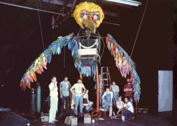 Seven college students sitting and standing in front of a big bird-looking puppet with yellow head feathers and colored wing feathers. 