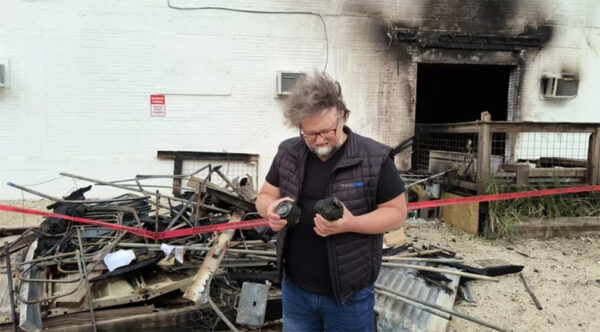 A photograph of a man standing outside of a fire damaged building. He looks at his hands as he holds damaged camera equipment.