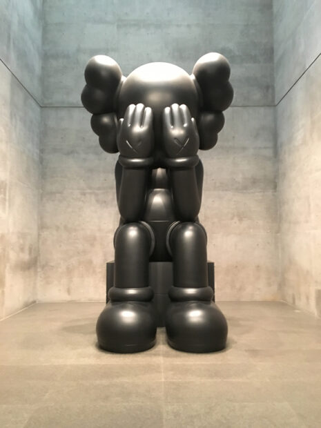 A large scale fiberglass and metal sculpture of a clown cartoon figure sitting with its hands over its eyes like it is crying.