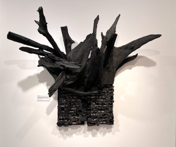 A large assemblage sculpture by Leonardo Drew. The piece consists of small pieces of wood stacked and glued together to create a rectangular form. Atop the form are large pieces of tree branches that jut from the top of the work and are also painted black.