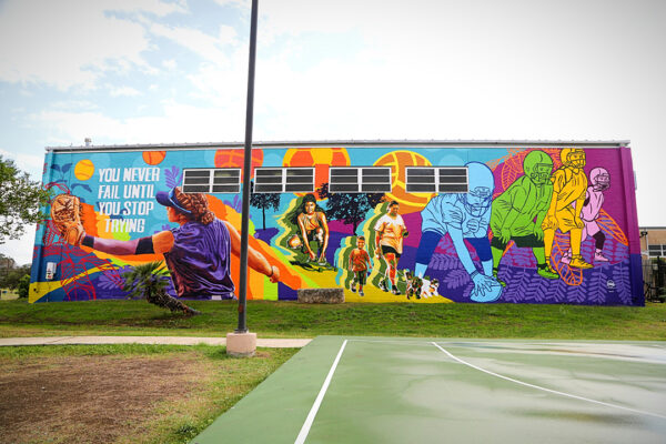 A large-scale mural on the exterior of a community center building. The mural uses vibrant colors and combines realistic portraiture with a flat graphic design style.