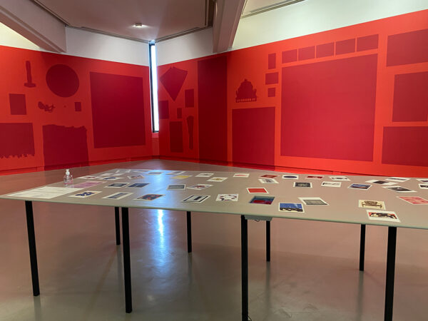 A gallery with two red walls, each wall has dark red silhouettes of objects that were not available to be hung in the gallery. In front of the wall is a table top with images of each of the objects for visitors to look at and guess where the object was supposed to hang.