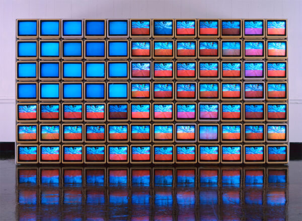 A wall of 84 small television sets. Together the installation appears to mimic the United States flag with a square of blue in the top left corner and stripes of red and blue across the rest of the piece.
