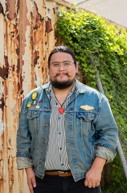 A photograph of writer and curator Reyes Ramirez. He stands outdoors and wears a jean jacket with pins in the upper right corner.