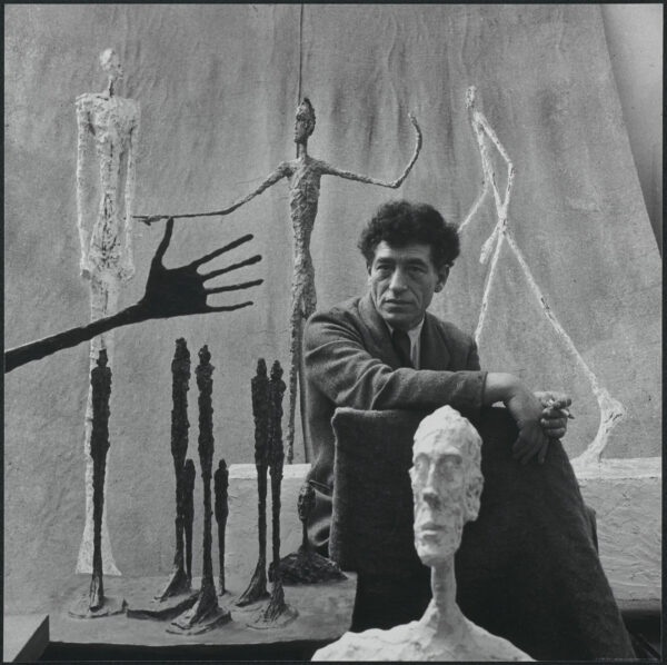A black and white portrait of Alberto Giacometti in his studio surrounded by figurative sculptures.