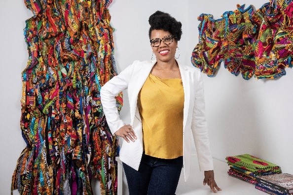 A photograph of artist Kathy Brown in her studio with brightly colored patterned fabrics hanging on the wall behind her.