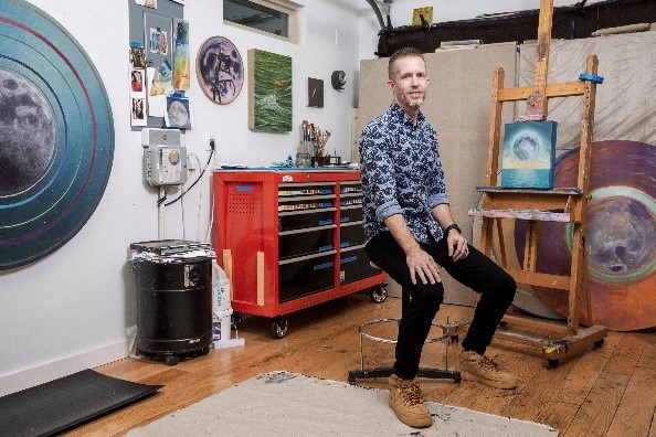 A photograph of artist Adam Fung in his studio with with various paintings of the moon and outer space on the walls behind him.