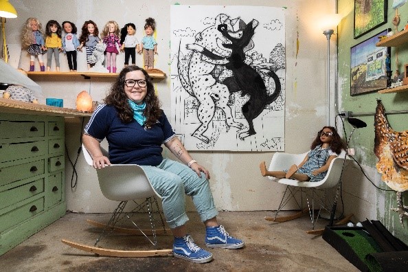 A photograph of Olivia Garcia-Hassell in her studio with various dolls standing on a shelf behind her.