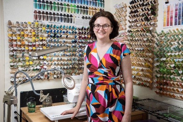 A photograph of artist Rebecca Shewmaker in her studio with a colorful wall of thread displayed behind her.