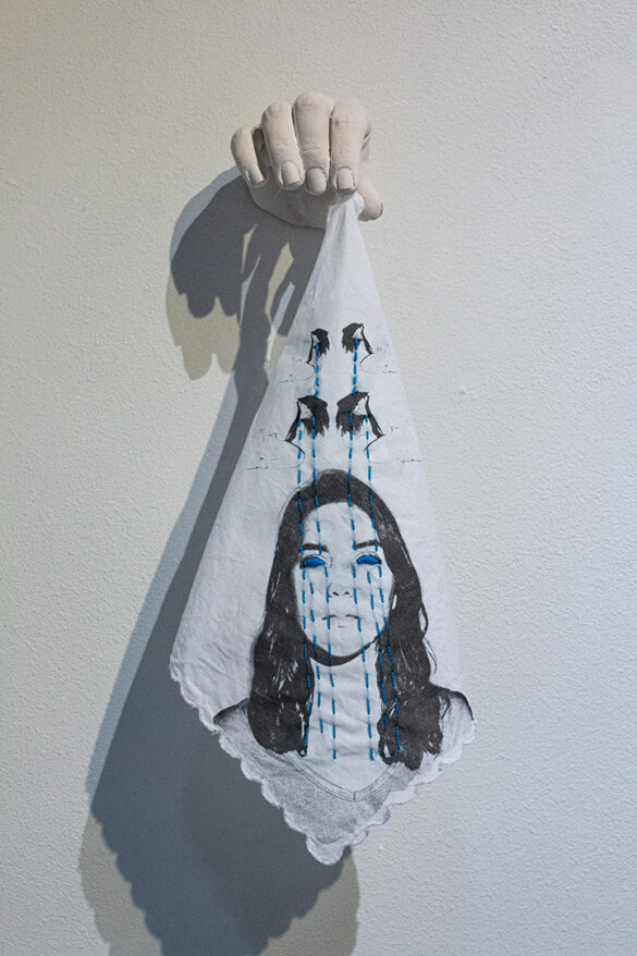 A work of art by Chantal Lesley. The work includes a plaster hand protruding from the wall and holding a handkerchief with a printed photograph of a woman and embroidered blue lines that look like rain and teardrops.