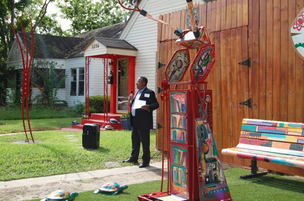 A photograph of a man speaking to a group that is out of sight of the camera. The man stands amongst colorful sculptural artworks.