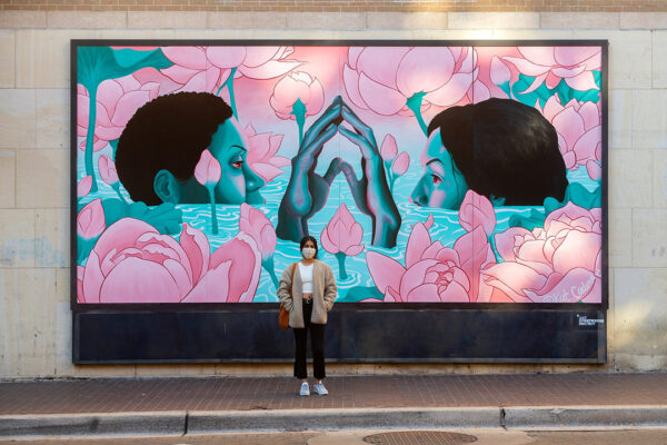 A photograph of artist Kat Cadena standing in front of a large-scale mural of two figures submerged in water, looking at each other above the surface and touching hands.
