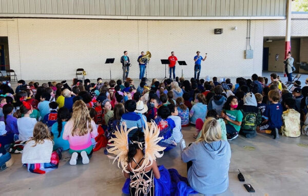A photograph of four bass instrumentalists playing for a large crowd of seated children.