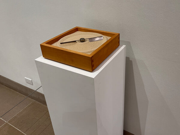 A photograph of a small sculpture by Mona Hatoum. The sculpture is a square wooden box filled with a sand. The center of the box has a blade which spins creating a circle in the sand.