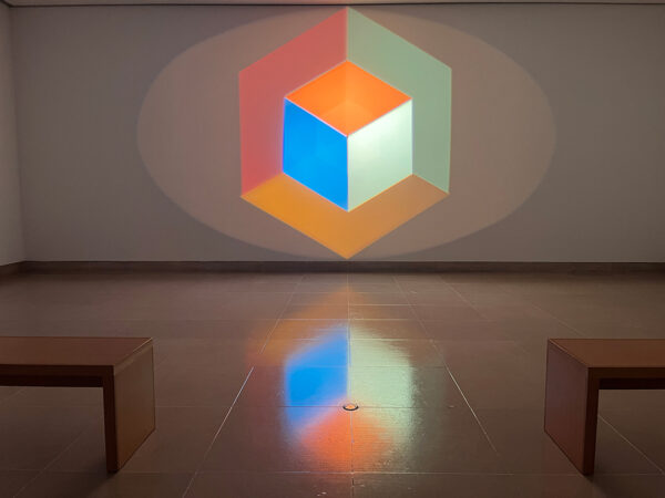 A photograph of a light projection on a wall. The projection creates two cubes made of various colors.
