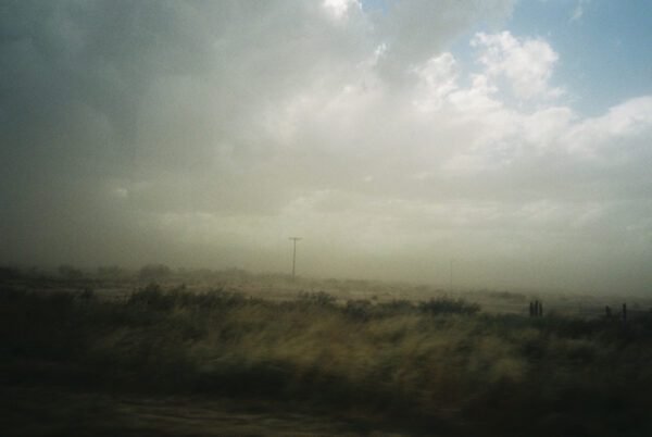 Fotografía borrosa de un paisaje desértico bajo un cielo cubierto de nubes. La hierba alta se inclina hacia la derecha por la fuerza de las ráfagas que también hacen volar polvo, tiñendo el cielo de un tono amarillento.