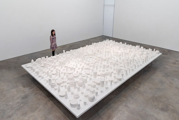 A photograph of a large-scale installation by Reynier Leyva Novo featuring a large rectangular table filled with an array of white sculptures of household items like vases and bowls.