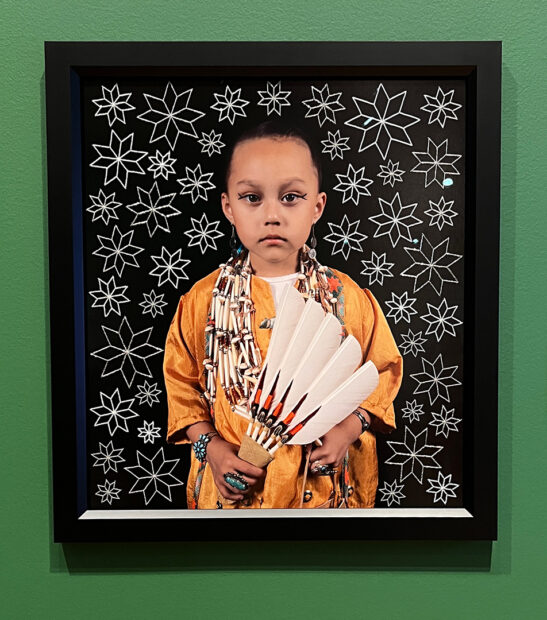 A photograph of a young child by Tom Jones. The black background behind the child is filled with white flower shapes created from small beads.