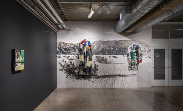 Installation view of work on a black wall and multicolored suits hanging in the space