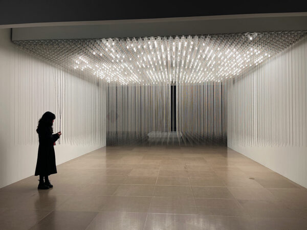A photograph of a light installation by Valeska Soares. The installation features thousands of simple light fixtures arranged in a grid on the ceiling. Each fixture has a long metal ball chain that can be pulled to turn the light off or on.