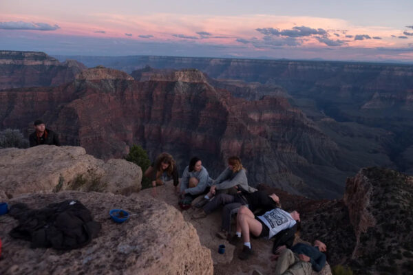 Un grupo de jóvenes, algunos sentados y otros acostados, están en el borde de un cañón. Detrás de ellos la luz del atardecer cae con tonalidades lilas.