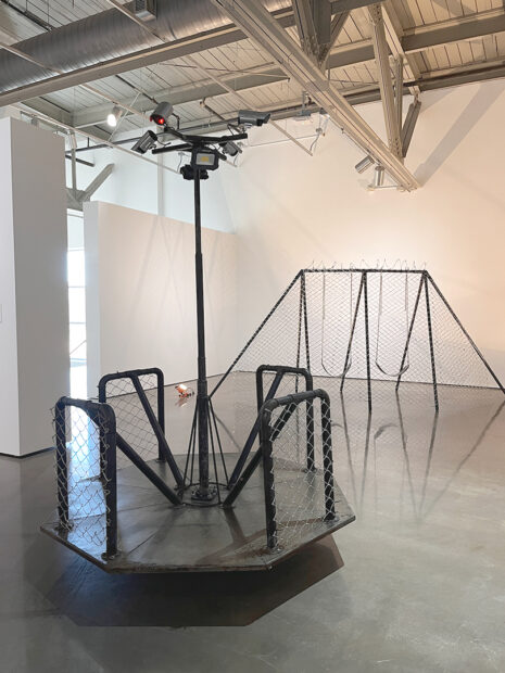 An installation image of a swingset and a child's merry-go-round altered to include chain-link fence, security cameras, and barbed wire.