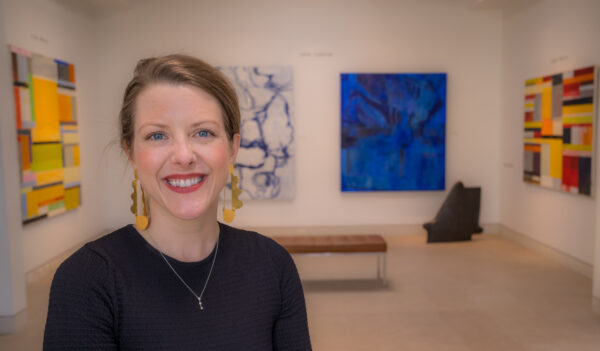 A photograph of Anne Kelly Lewis in a gallery with large abstract paintings behind her.